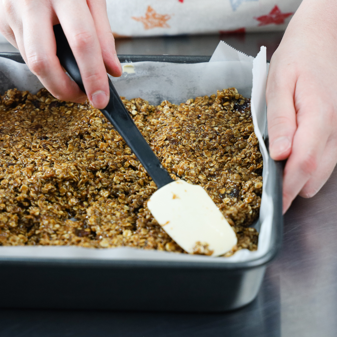 Sticky Toffee Flapjack being pressed into a square tin, using a white spatula.